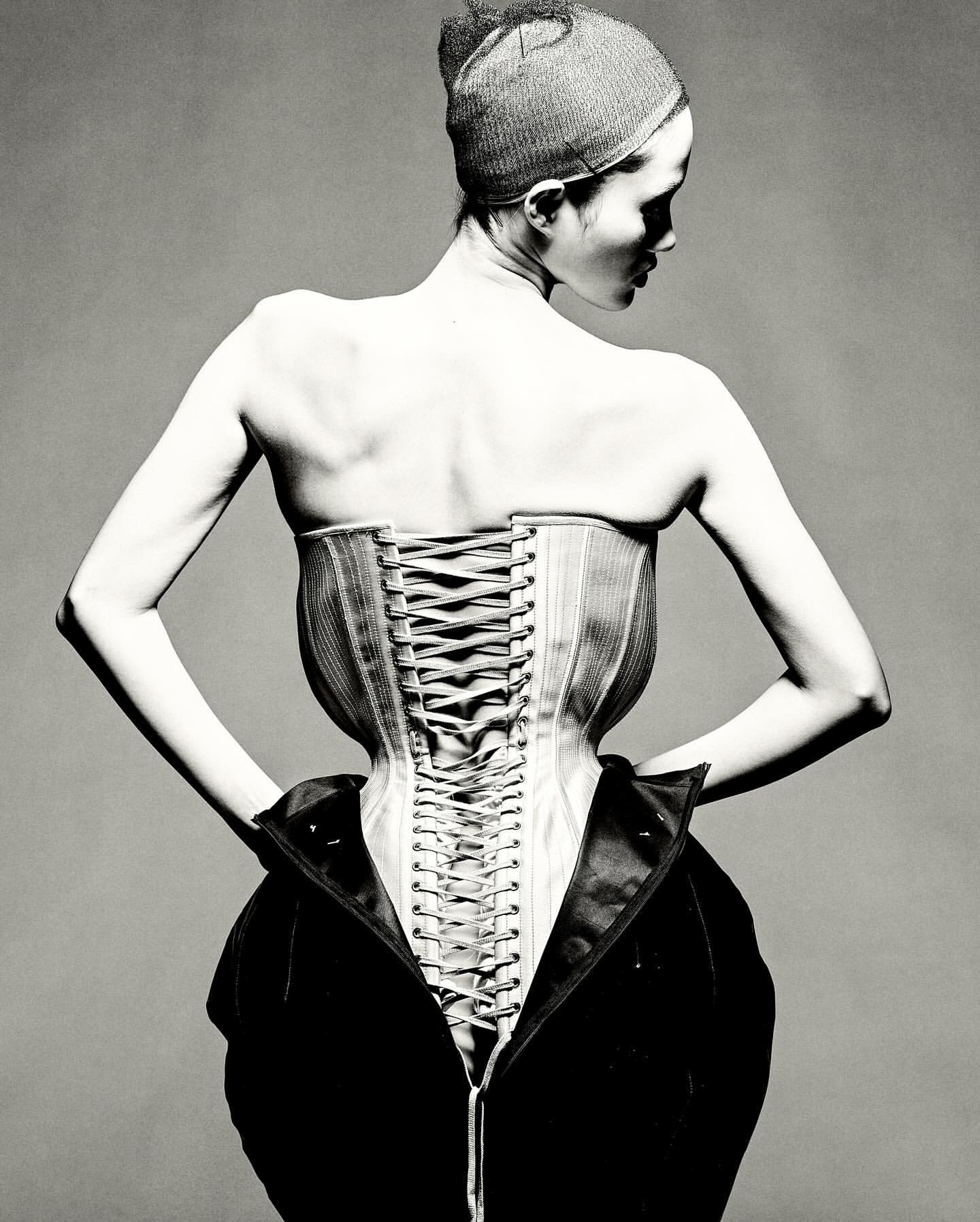 Monochrome back view of an adult female with a modern corset and visible lacing, accented by a partially unzipped dark coat.