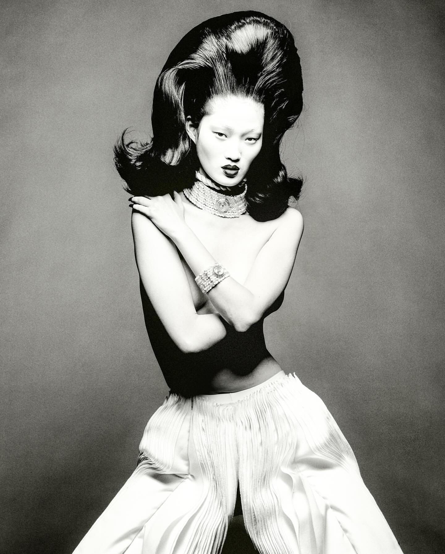 Monochrome fashion portrait of an Asian model with voluminous dark hair, statement jewelry, and white wide-leg pants, set against a grey backdrop.