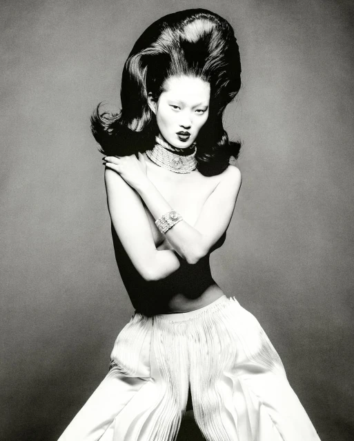 Monochrome fashion portrait of an Asian model with voluminous dark hair, statement jewelry, and white wide-leg pants, set against a grey backdrop.