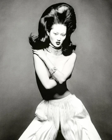 Monochrome fashion portrait of an Asian model with voluminous dark hair, statement jewelry, and white wide-leg pants, set against a grey backdrop.