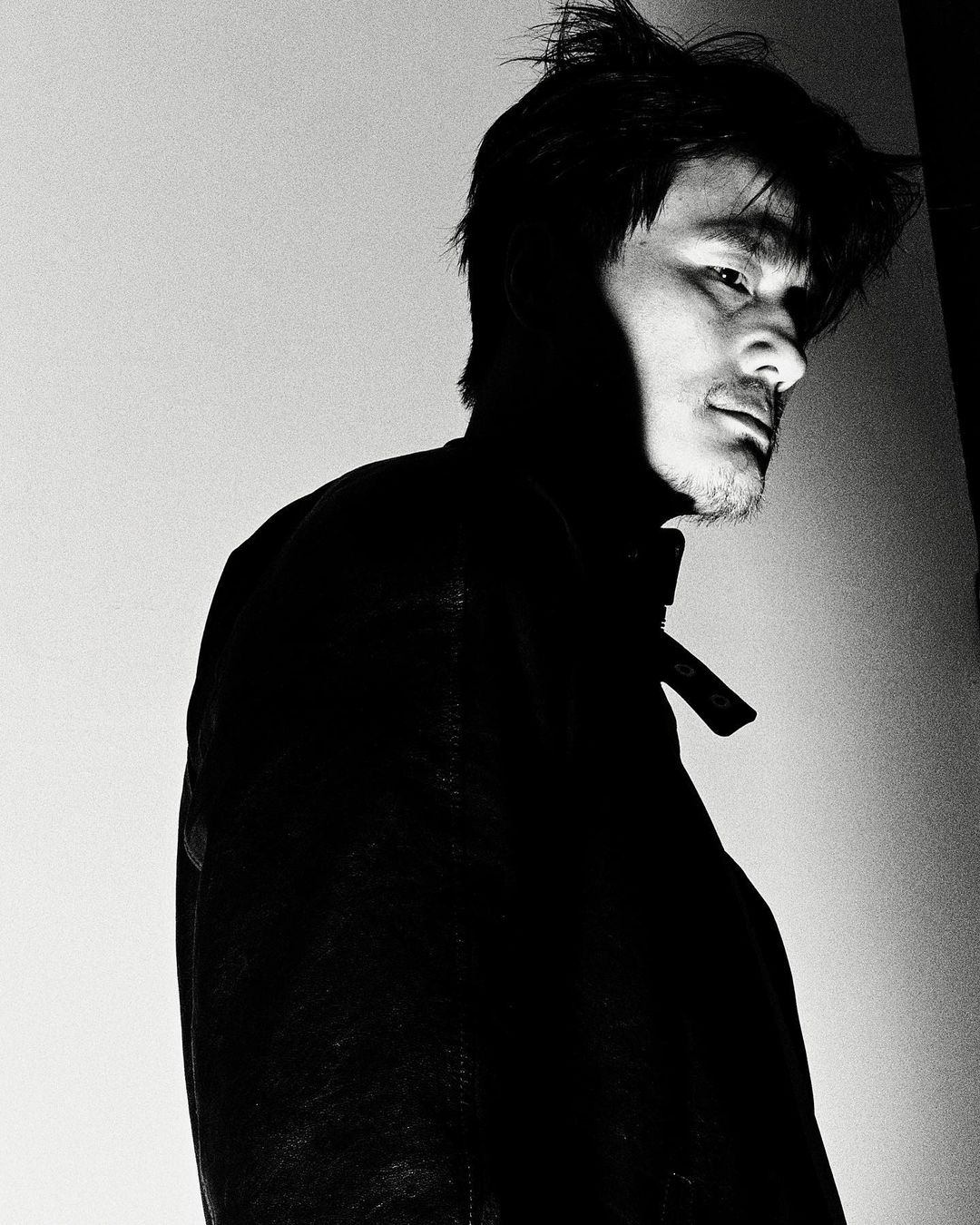 Monochrome portrait of a man in a jacket with dark shadows, showcasing a candid and intimate style with a simple background.
