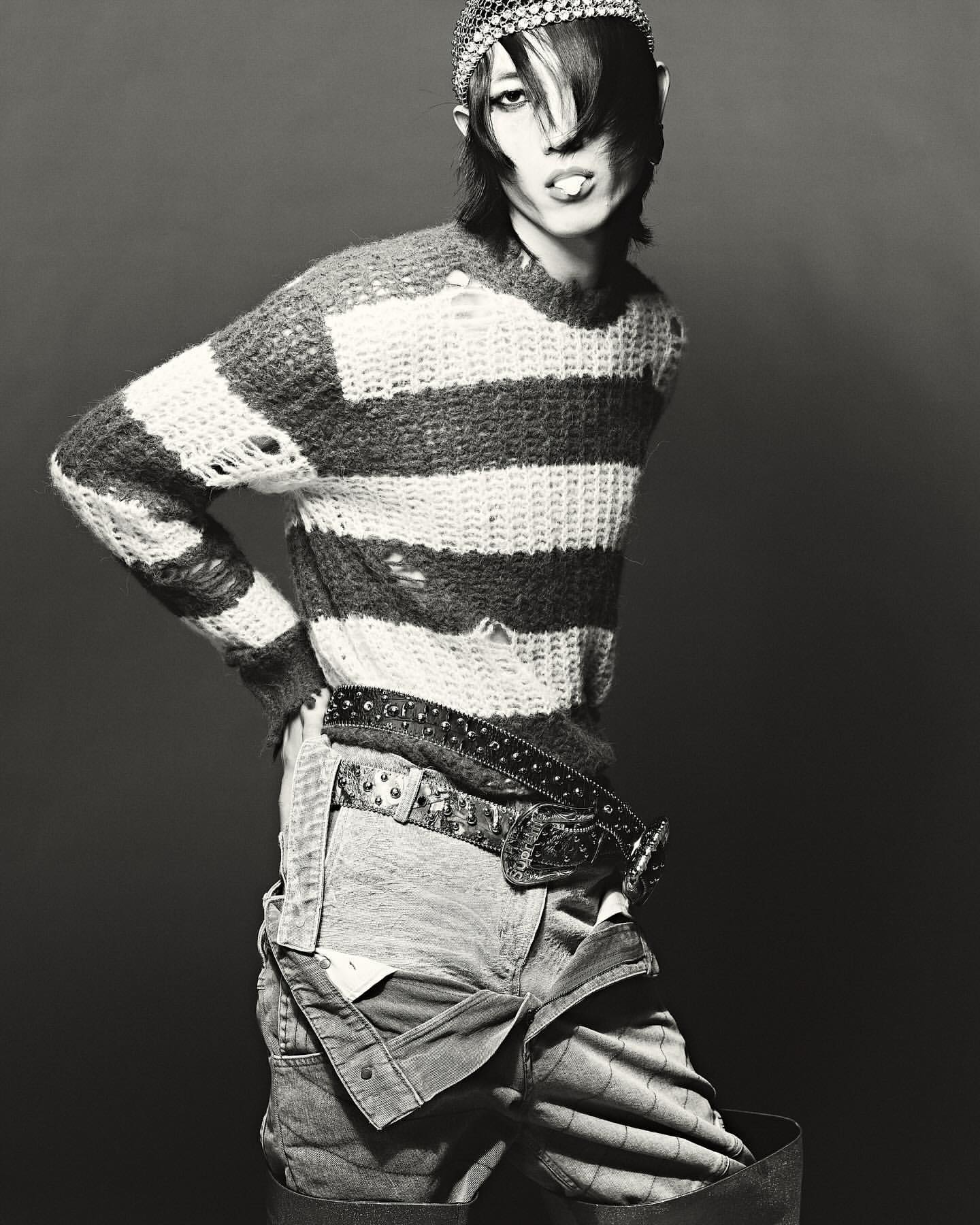 Striking black and white studio shot of androgynous person in distressed striped sweater and overalls, accessorized with studded belt and headpiece.