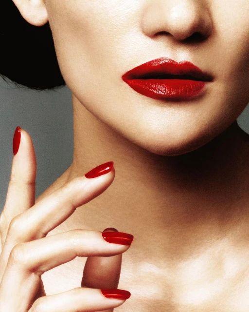 Close-up of a woman with red lipstick and matching manicure, highlighting beauty and fashion details against a neutral gray backdrop.