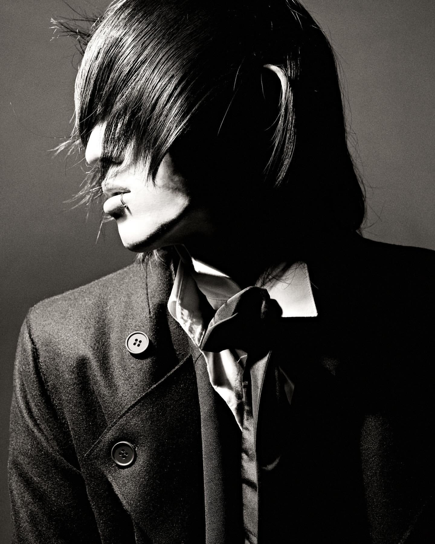 Monochrome portrait of an adult male with dark hair partially covering his face, wearing a collared shirt and a buttoned coat indoors.