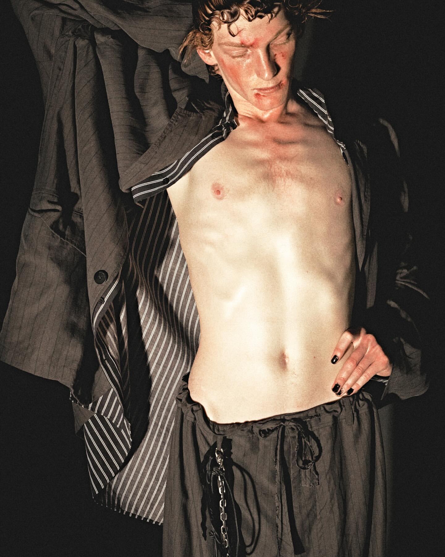 Shirtless adult male poses in dark studio with a dark pinstripe blazer. He wears dark drawstring trousers, and has a light complexion.
