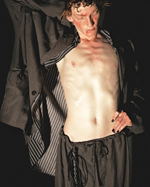 Shirtless adult male poses in dark studio with a dark pinstripe blazer. He wears dark drawstring trousers, and has a light complexion.