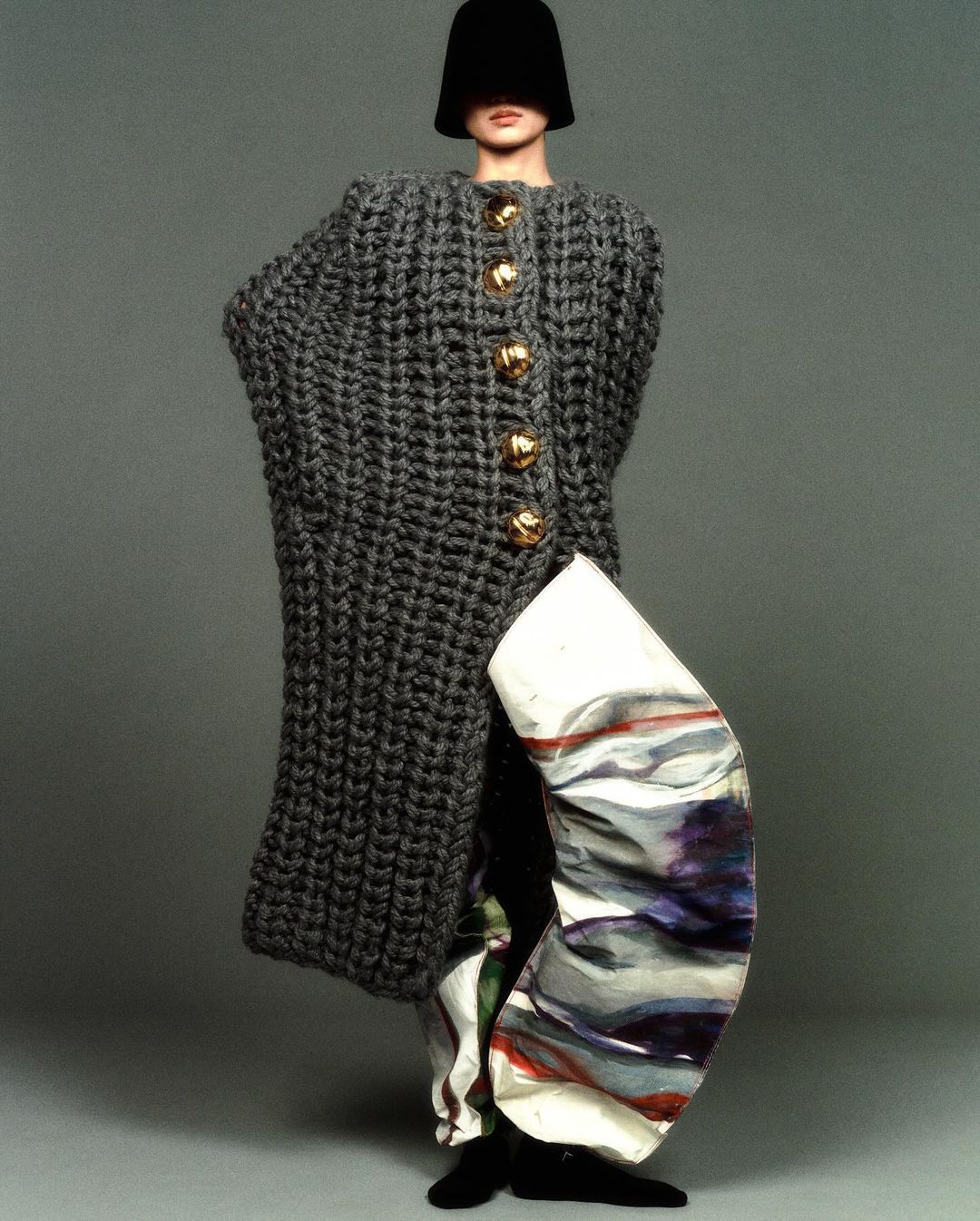 Fashion shot of a slender model wearing a knitted gray coat with gold buttons and abstract painted pants against a gray backdrop.