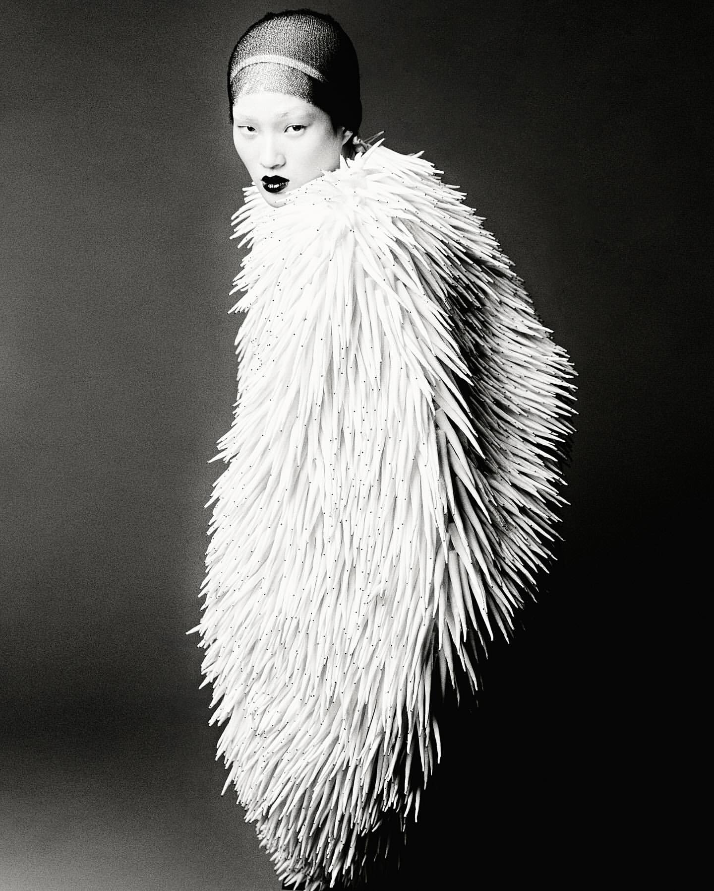 Black and white portrait of a woman wearing a textured white coat-like garment against a dark gray background in a studio setting.