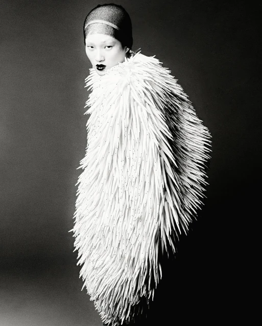 Black and white portrait of a woman wearing a textured white coat-like garment against a dark gray background in a studio setting.