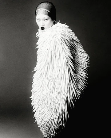 Black and white portrait of a woman wearing a textured white coat-like garment against a dark gray background in a studio setting.