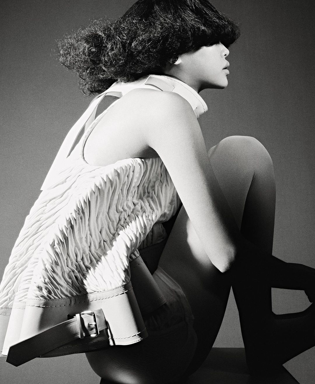 Monochrome studio portrait of a seated adult woman in a white textured top with dark hair partially covering her face, against a gray backdrop.