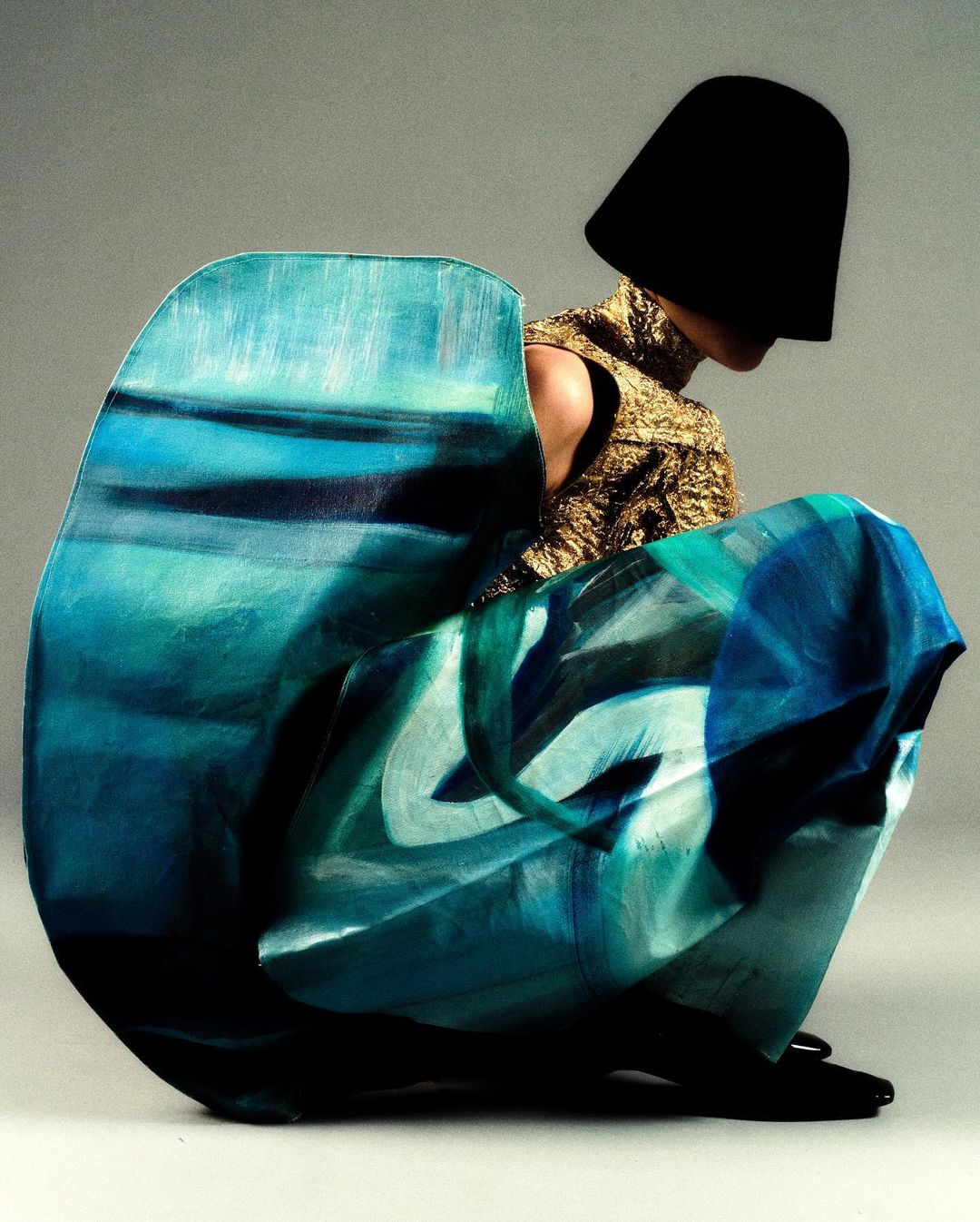 Fashion shot featuring a person in a gold top and large blue patterned fabric. Head is covered by a black hat-like object.