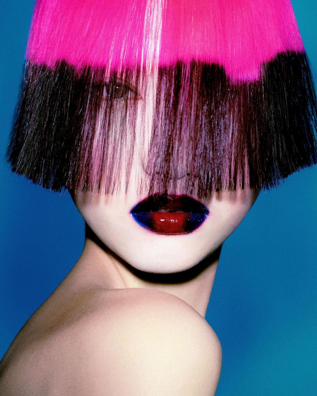 Headshot of a person with a pink and black bob haircut and a dark red and blue lip, set against a bright blue background.