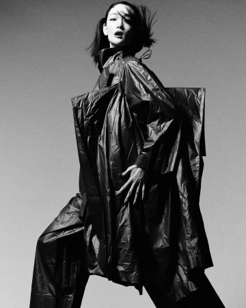 Monochrome fashion portrait of a model in an oversized dark sculptural coat against a grey studio backdrop