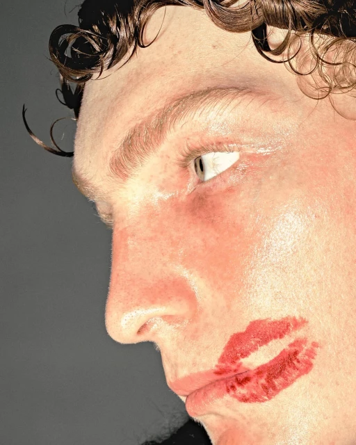 Close-up portrait of a man's face with a red lipstick kiss on his cheek, curly hair visible, against a solid grey background.