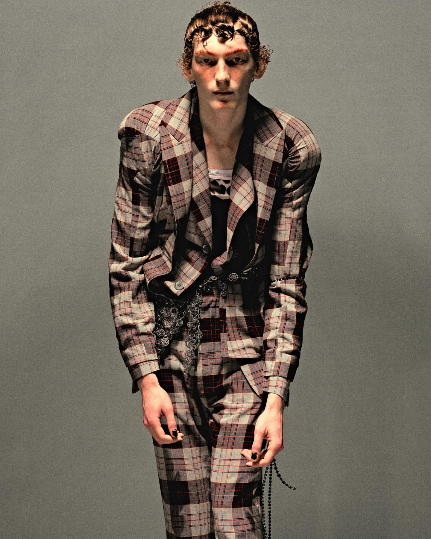 Fashion portrait of a model wearing a plaid suit. The background is grey. The subject is posed in a full-body shot indoors.