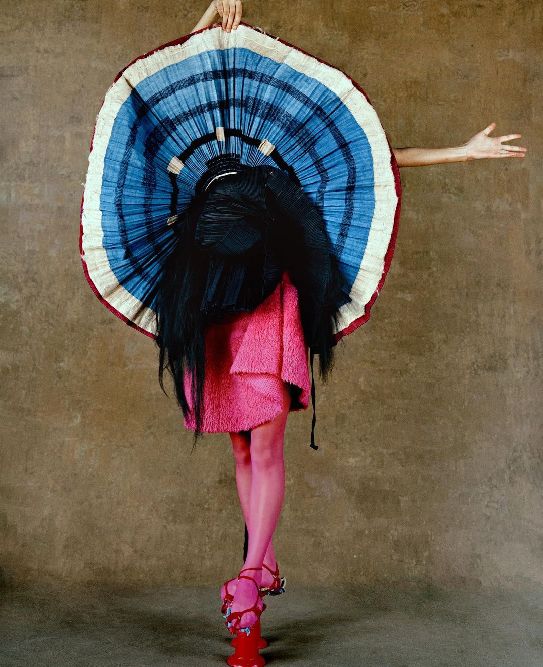 Person wearing oversized blue and white striped disc-shaped headpiece and pink dress standing on a red object in a studio setting.