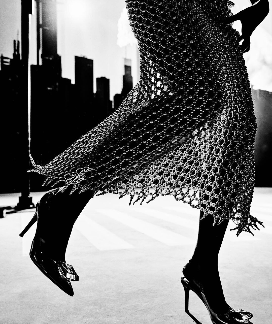 Fashionable woman strides forward, showcasing a distinctive dress with a net-like design, paired with high heels, against a cityscape backdrop.