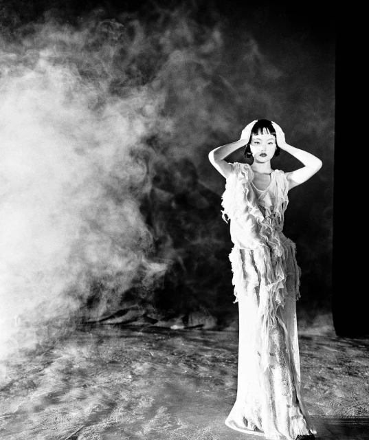 Black and white studio portrait of a woman in textured dress with smoke effect, hands on head, medium shot, creating dramatic fashion visual.