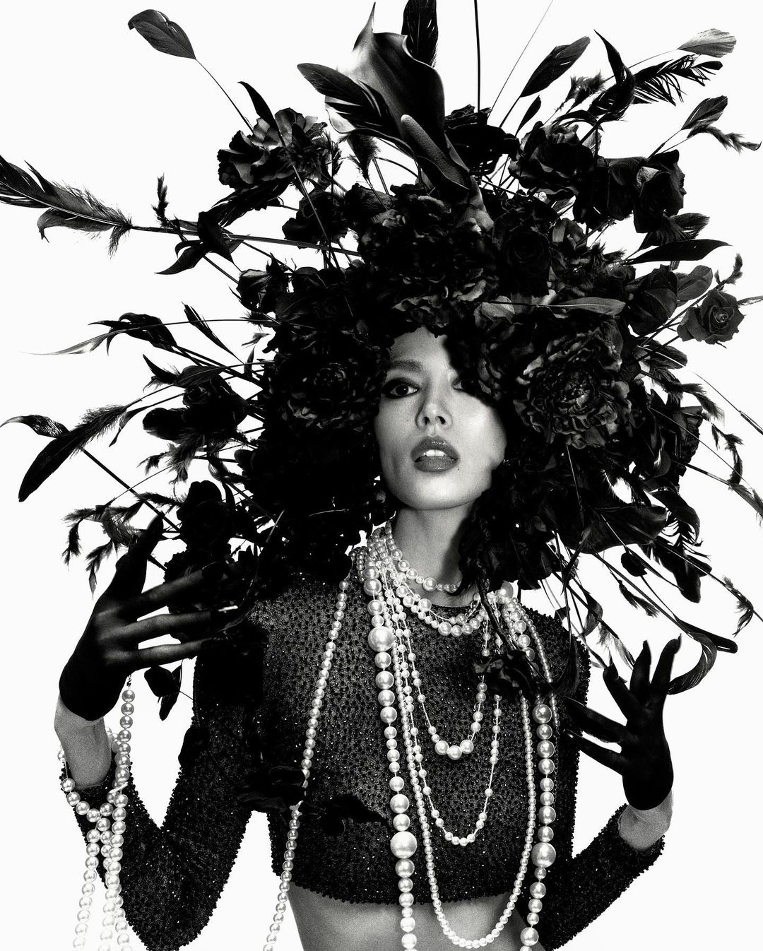 Striking black and white portrait of a woman adorned with a large floral headdress and layers of pearl necklaces in a studio setting.