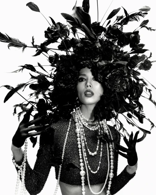 Striking black and white portrait of a woman adorned with a large floral headdress and layers of pearl necklaces in a studio setting.
