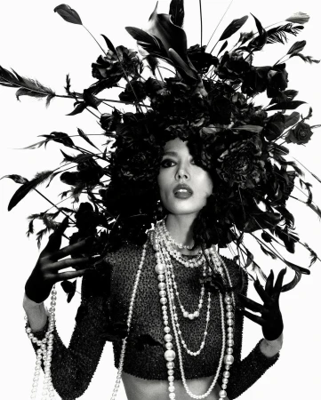 Striking black and white portrait of a woman adorned with a large floral headdress and layers of pearl necklaces in a studio setting.