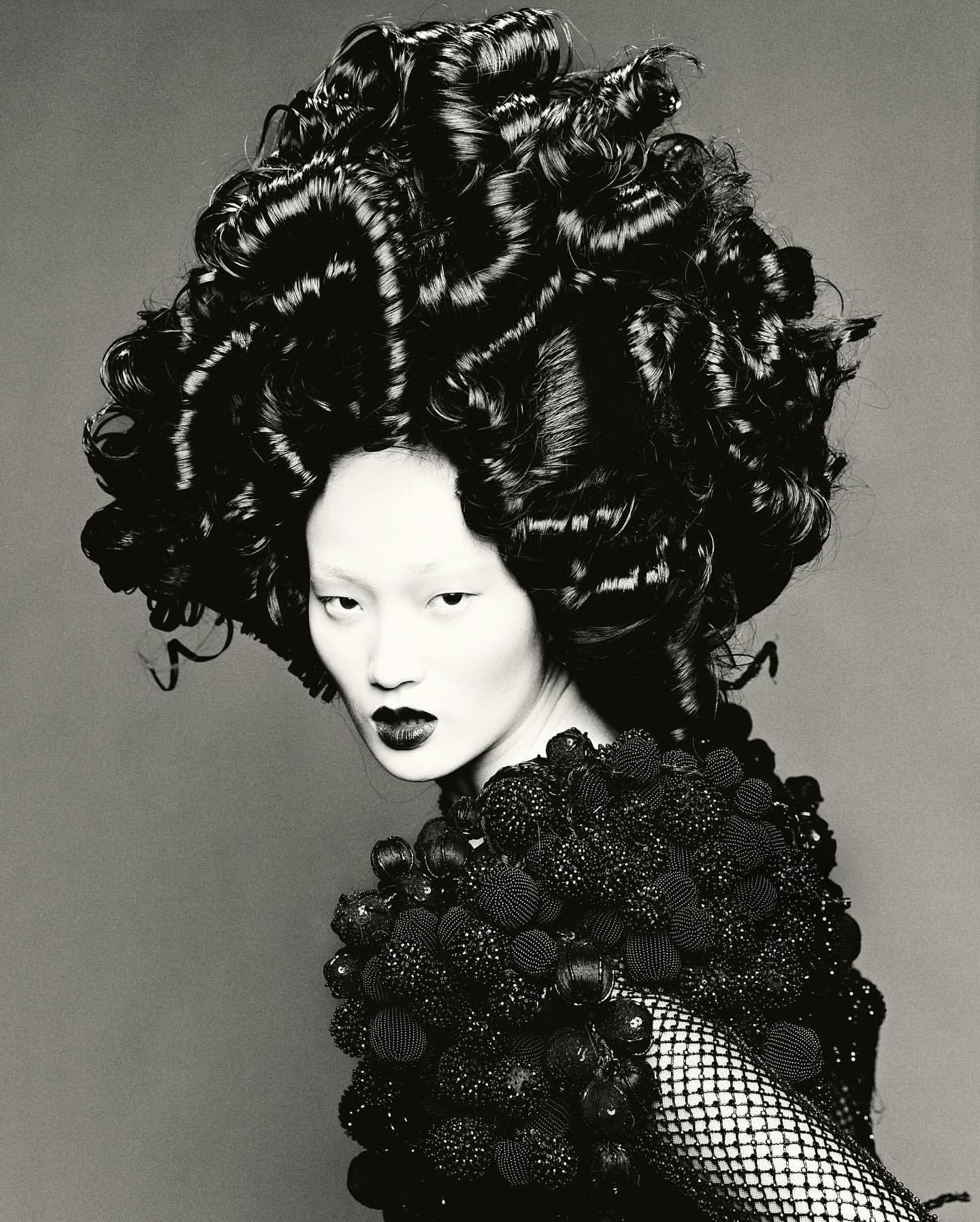 Asian woman with avant-garde curly hairstyle and decorative shoulder piece posing against a gray background in a studio lighting.