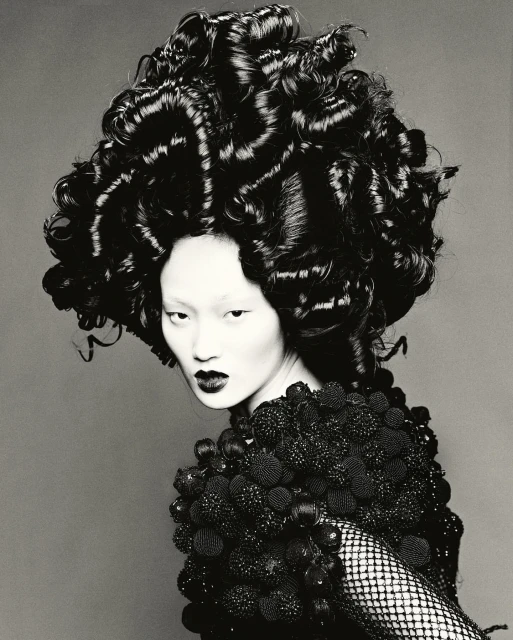Asian woman with avant-garde curly hairstyle and decorative shoulder piece posing against a gray background in a studio lighting.
