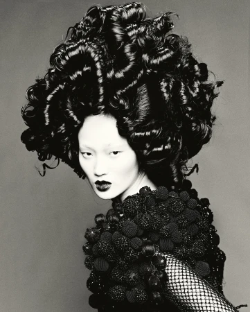 Asian woman with avant-garde curly hairstyle and decorative shoulder piece posing against a gray background in a studio lighting.