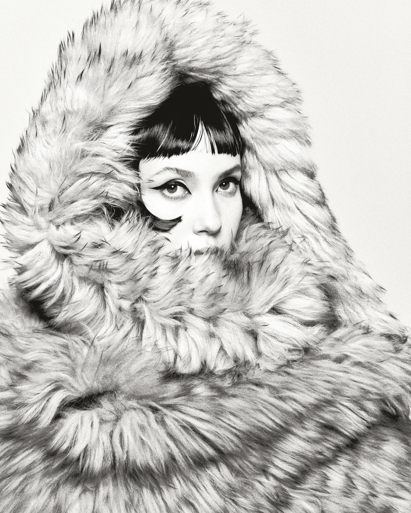 Portrait of a woman with short black hair and heavy eyeliner, wearing a large fur hood and coat against a neutral background.