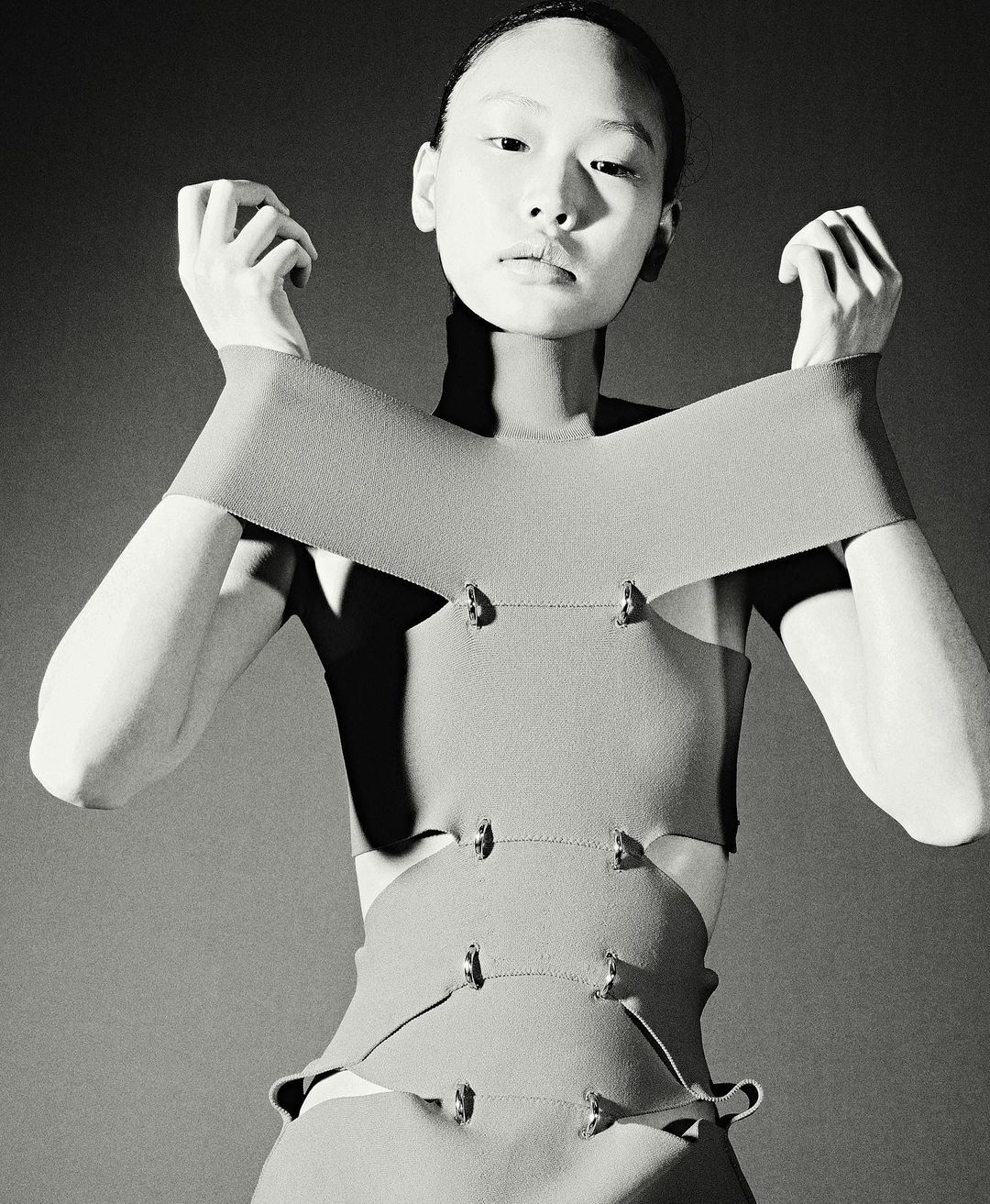 A monochrome portrait of an Asian woman in a sleeveless top with metal details, posing with hands near her face in a photo studio.