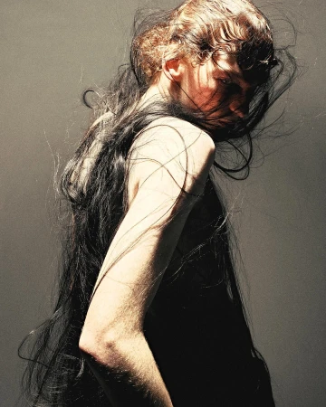 Model with long dark hair blowing across face in profile wearing a black top against a dark grey textured background