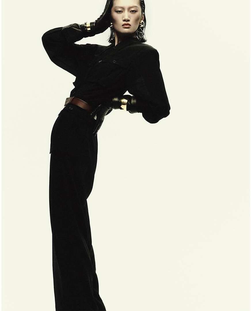 Fashion shot of an Asian woman wearing black clothing and gold bracelets, leaning back against a light background in a studio.