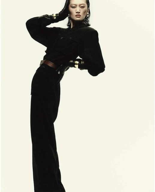Fashion shot of an Asian woman wearing black clothing and gold bracelets, leaning back against a light background in a studio.