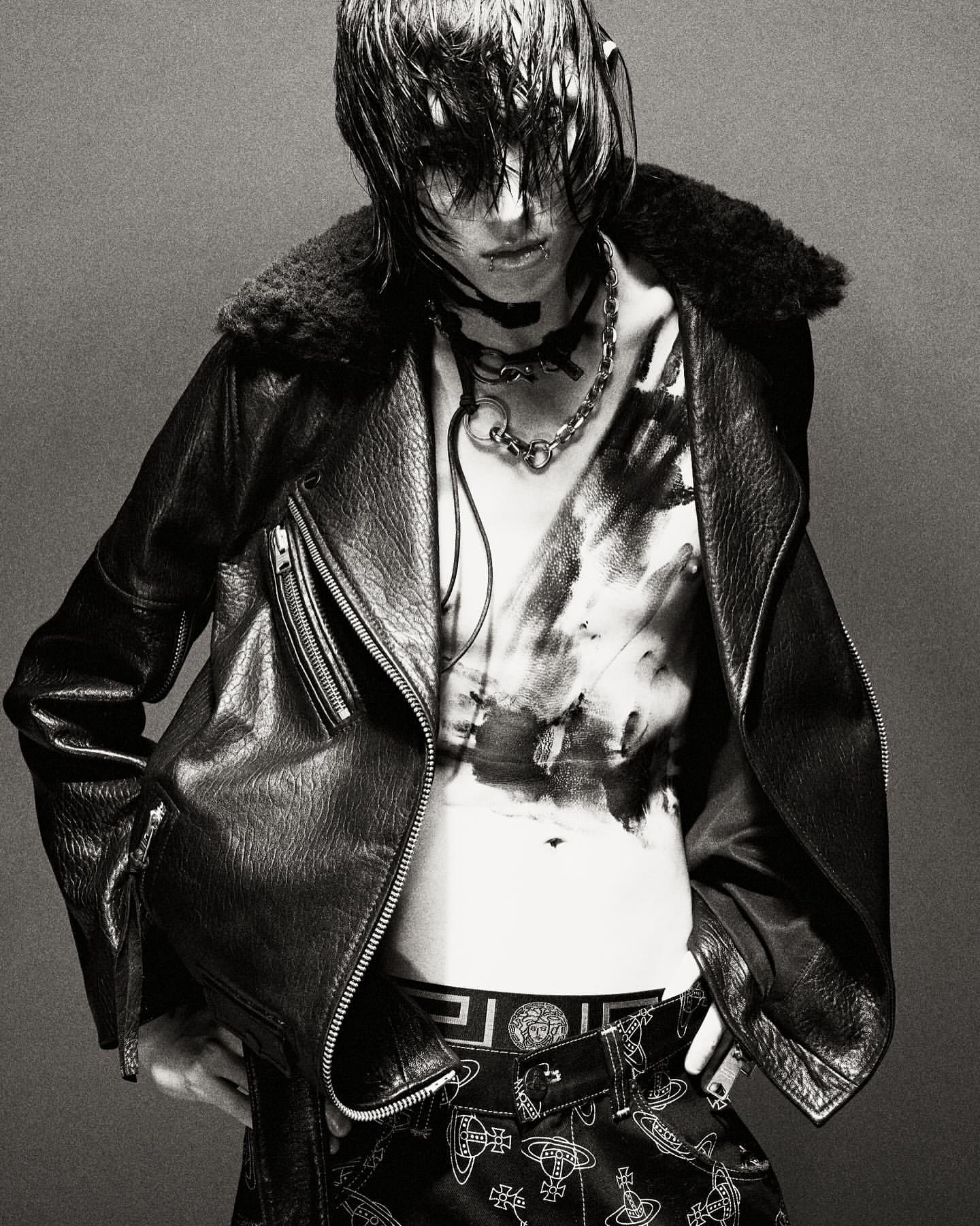A person wearing a leather jacket with fur collar and designer-patterned pants poses in a studio with a dark gray backdrop.