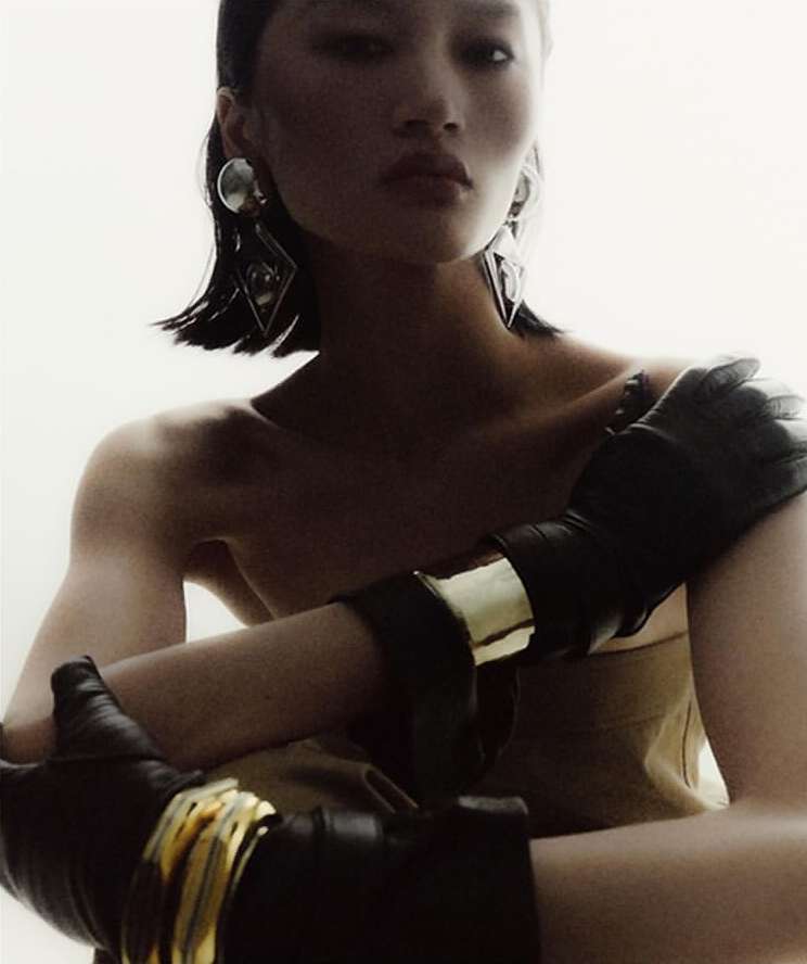 Model with dark wet-look hair wearing geometric silver earrings and black leather gloves with gold bracelets against a bright white backdrop