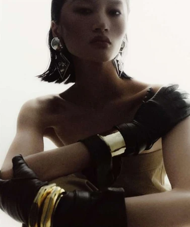 Model with dark wet-look hair wearing geometric silver earrings and black leather gloves with gold bracelets against a bright white backdrop