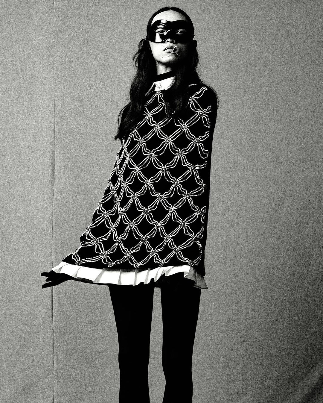 Black and white fashion shot of a woman wearing a patterned top, leggings, and a mask, set against a plain backdrop in a studio environment.