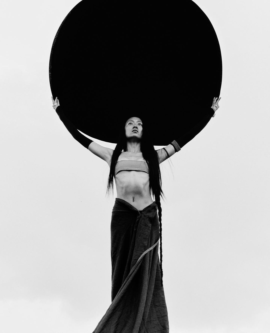 Artistic black-and-white portrait of a slender woman holding a large black disc overhead in an open, bright outdoor environment.