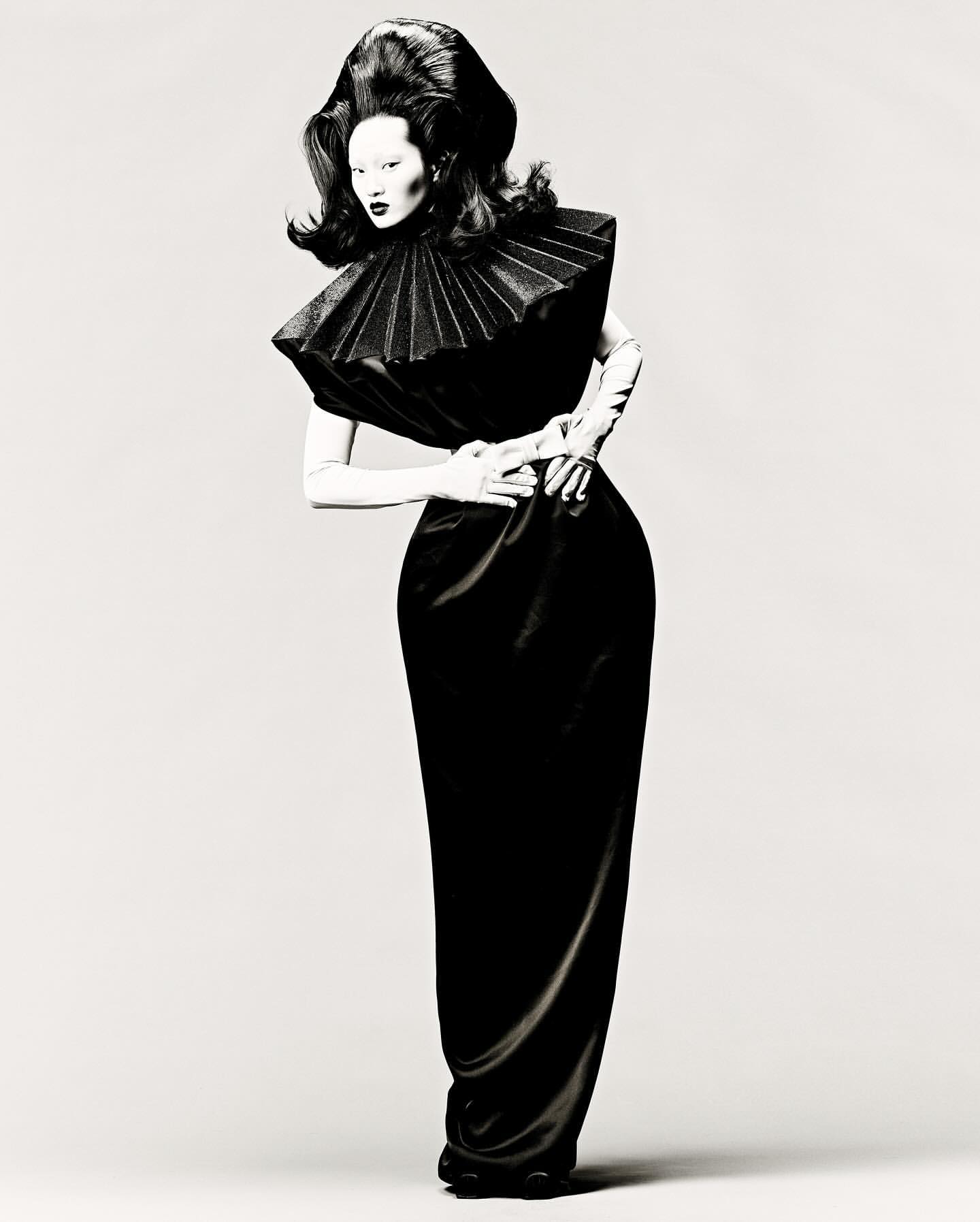 Woman with dramatic hair and black gown poses against a plain background in a fashion-editorial style full shot.