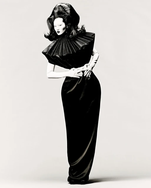 Woman with dramatic hair and black gown poses against a plain background in a fashion-editorial style full shot.