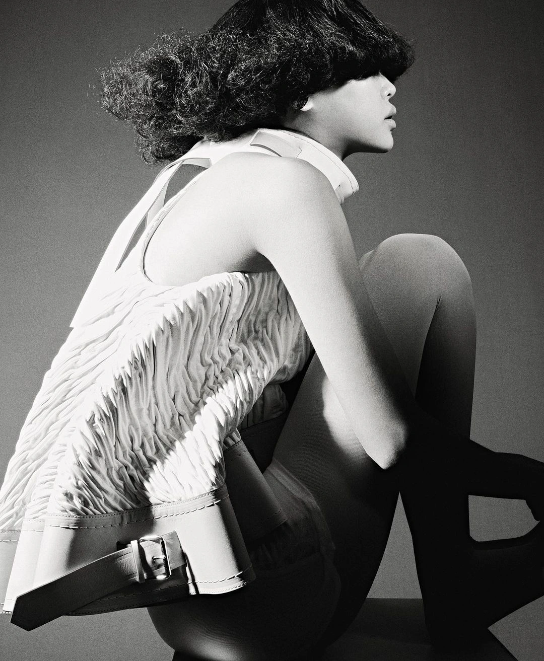 Monochrome studio portrait of a model with textured dark hair seated in profile wearing a textured white sleeveless top
