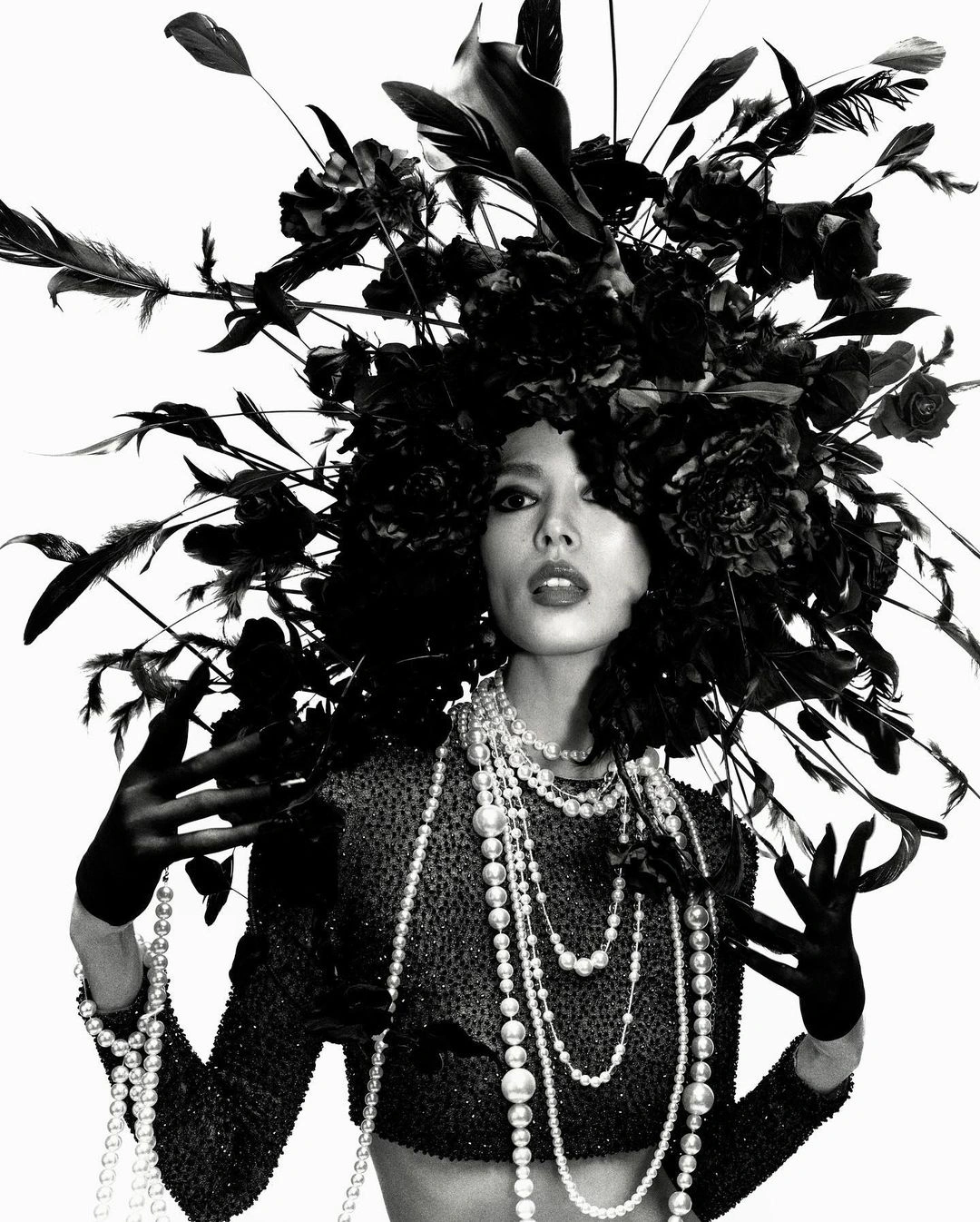 Model wearing a massive black floral headpiece and layered pearl necklaces against a bright white background