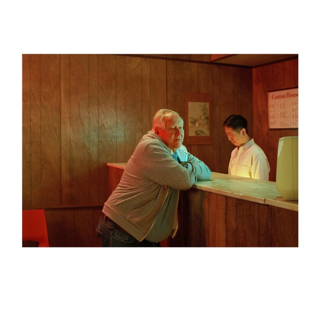An older adult male leans on a counter in a restaurant with an Asian male employee in the background. Wood paneling walls.