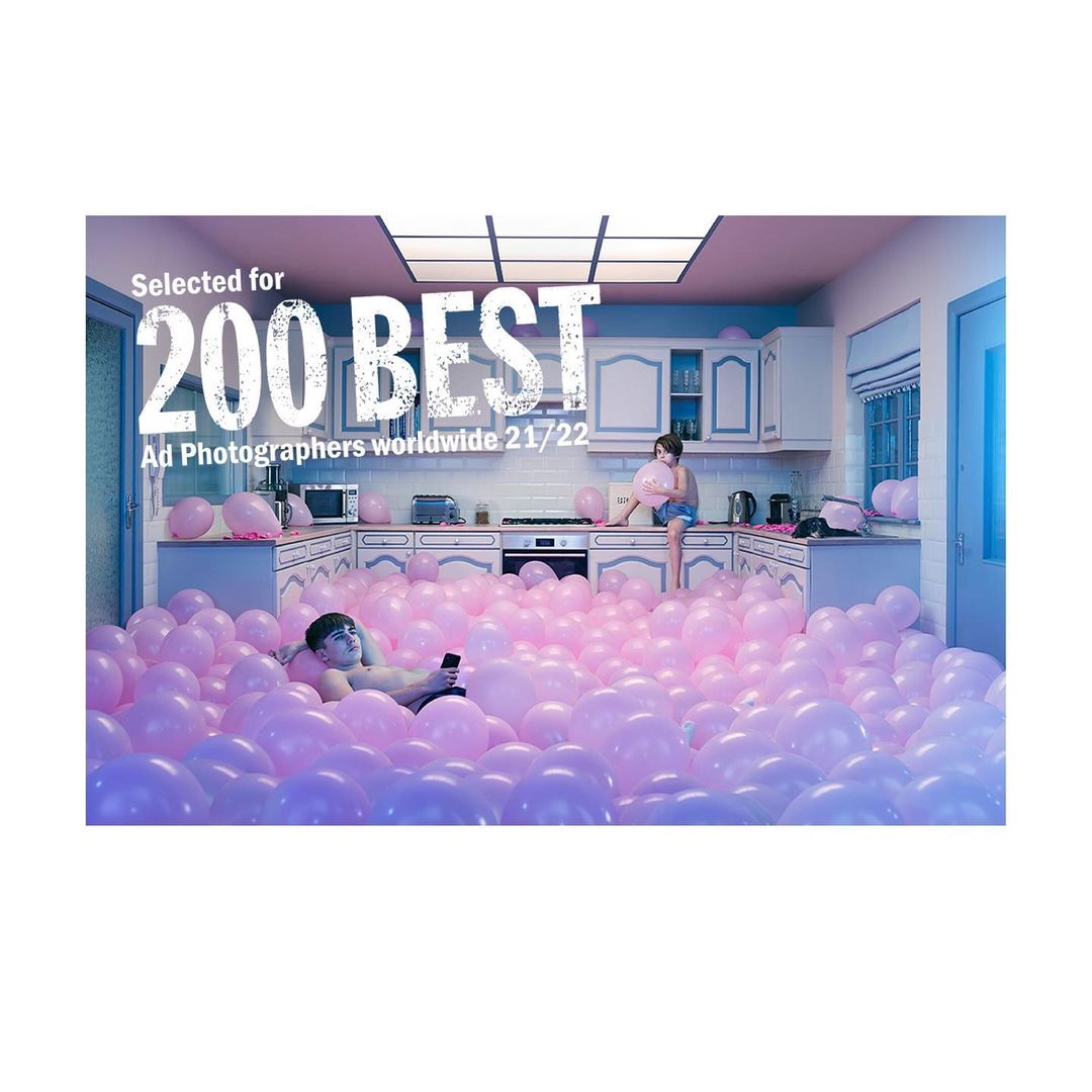 Man lounging in a kitchen filled with pink balloons and a child sitting on the counter holding a pink balloon