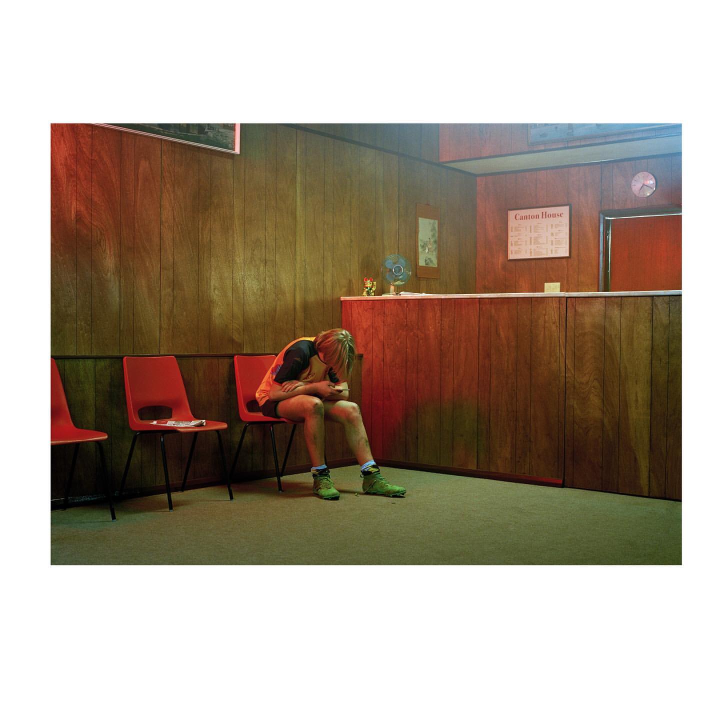 A person sits alone in a waiting room, head in their hands, near red chairs and wood-paneled walls, creating a somber indoor scene.