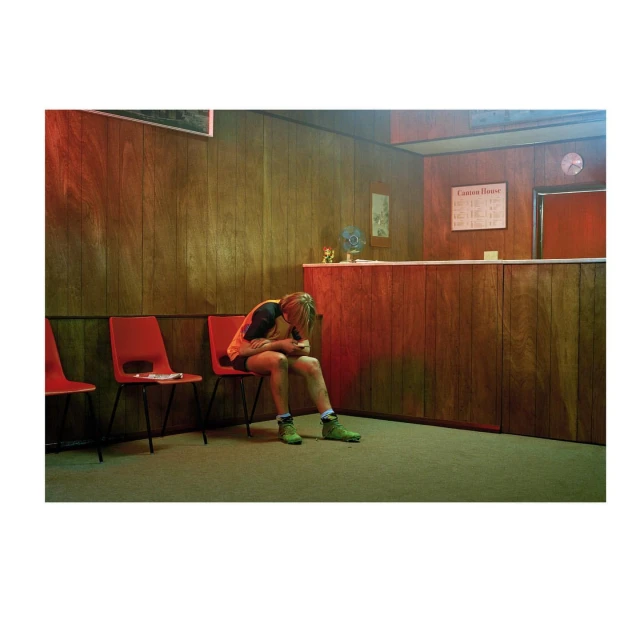 A person sits alone in a waiting room, head in their hands, near red chairs and wood-paneled walls, creating a somber indoor scene.