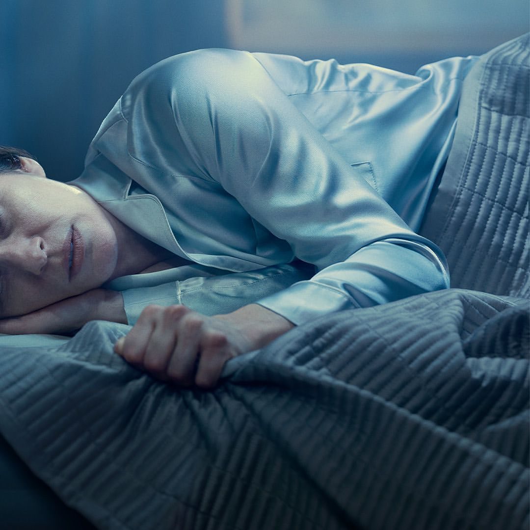 Adult woman sleeps in bed under a gray blanket, wearing a satin pajama top in an indoor setting with a blue color grading.