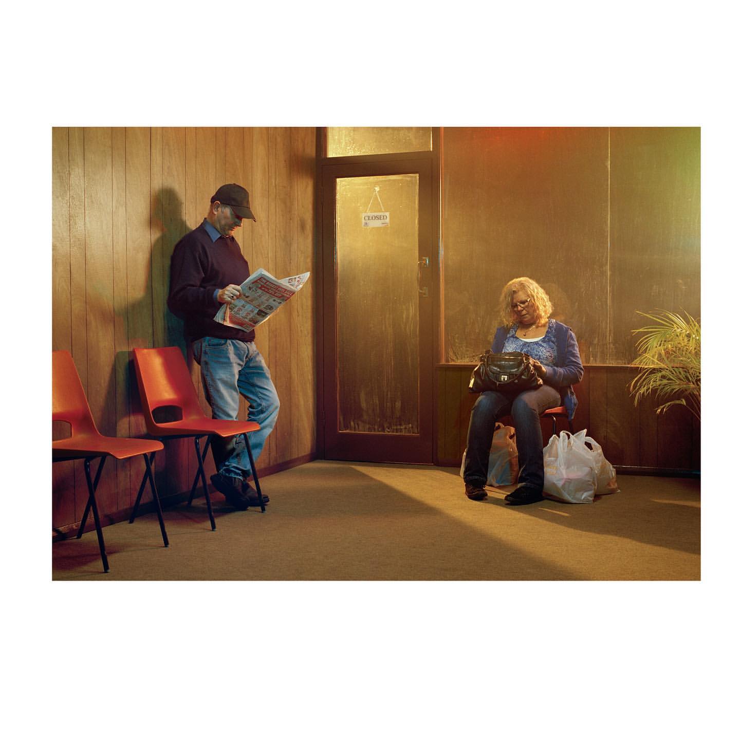 Indoor waiting area with a man reading newspaper and a woman with purse sitting next to shopping bag, warm, yellow lighting.