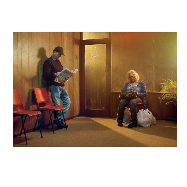 Indoor waiting area with a man reading newspaper and a woman with purse sitting next to shopping bag, warm, yellow lighting.